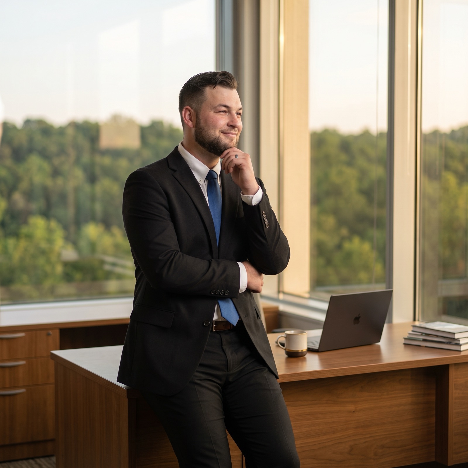 Valentin Gorobets in a black suit and blue tie, leaning on a walnut desk beside a tall window, smiling out at green hills